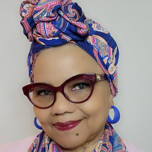 May include: A woman wearing a blue paisley head wrap and matching scarf. She is also wearing burgundy cat-eye glasses and blue hoop earrings. The head wrap and scarf feature a pattern of pink, orange, and white accents.