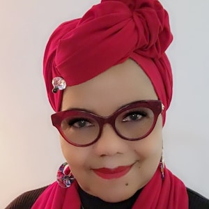 Op de afbeelding: Een vrouw draagt een rode turban met een juweelaccent, een rode bril, rode oorbellen, een rode sjaal en een zilveren ketting met een hartvormige hanger.