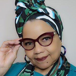 May include: A woman wearing a teal blue shirt with a black, green and white patterned scarf. She is wearing a pair of red cat-eye glasses and has a silver heart pendant necklace.