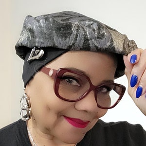 Könnte beinhalten: Eine Frau trägt eine schwarze und silberne gemusterte Baskenmütze mit einer silbernen Nadel, eine rot umrahmte Brille, eine silberne Kettenkette mit einem Herzanhänger und silberne Kettenohrringe. Sie trägt ein schwarzes Hemd.