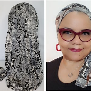 Puede incluir: Un turbante de tela de seda con estampado de serpiente en blanco y negro. El turbante está anudado en la parte superior de la cabeza y los extremos están colgando a los lados del cuello.