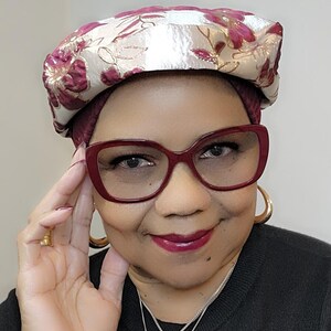 Könnte beinhalten: Eine Frau trägt eine burgunderrote und goldene, floral gemusterte Baskenmütze mit weißem Satin-Futter. Sie trägt außerdem eine rot umrandete Brille und ein schwarzes Hemd.