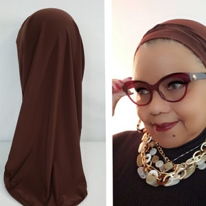 May include: A brown jersey knit turban head wrap. The turban is shown on a mannequin head and on a person wearing it. The person is wearing a black turtleneck sweater and a gold chain necklace with white beads.