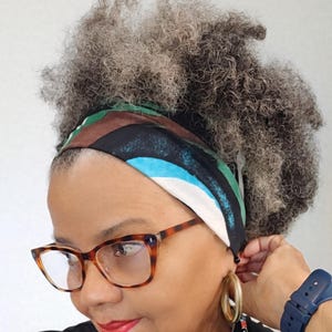 Peut inclure: Un foulard en soie coloré avec un motif géométrique bleu, blanc, marron et noir est porté en bandeau. La personne porte des lunettes écailles et des créoles dorées. Une montre bleue est au poignet.