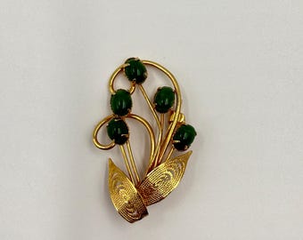 May include: A gold-toned brooch with a floral design featuring five green cabochon stones. The brooch has a delicate, intricate design with openwork details.