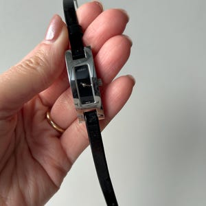 Puede incluir: Un reloj de pulsera rectangular plateado con esfera negra y correa de cuero negro. La esfera del reloj tiene la palabra "Gucci" impresa. El reloj se sostiene en una mano.