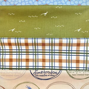 May include: A close-up of a fabric with a dinosaur theme. The fabric is beige with brown, green, and blue accents. The fabric features illustrations of dinosaurs with their names and descriptions. The fabric is layered on top of a green fabric with white dinosaur footprints and a blue and white fabric with a stone pattern.