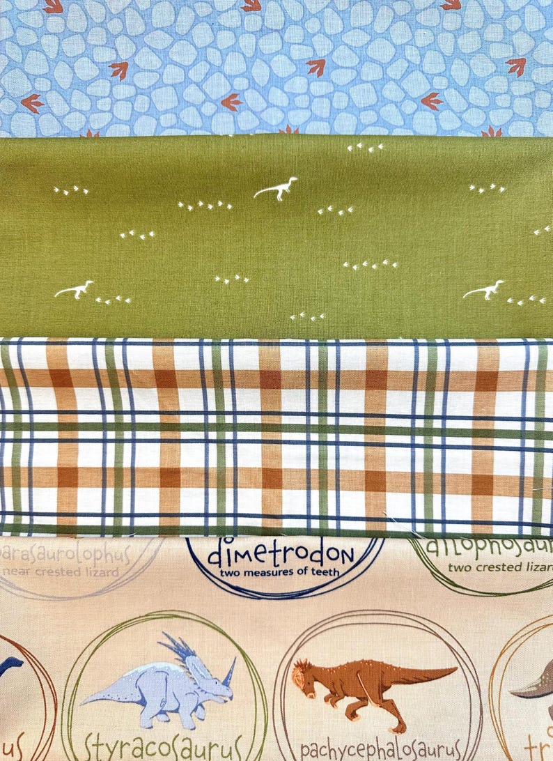 May include: A close-up of a fabric with a dinosaur theme. The fabric is beige with brown, green, and blue accents. The fabric features illustrations of dinosaurs with their names and descriptions. The fabric is layered on top of a green fabric with white dinosaur footprints and a blue and white fabric with a stone pattern.