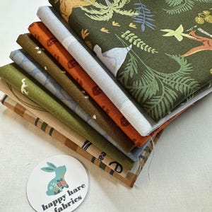May include: A stack of six fabric squares with different patterns, including a green fabric with a jungle animal print, a blue fabric with a white animal print, and a brown fabric with a white footprint print. The fabric squares are stacked on top of each other and have a white background. The bottom fabric square has a brown and white striped pattern. A white circle with the text "happy hare fabrics" is in the lower left corner of the image.