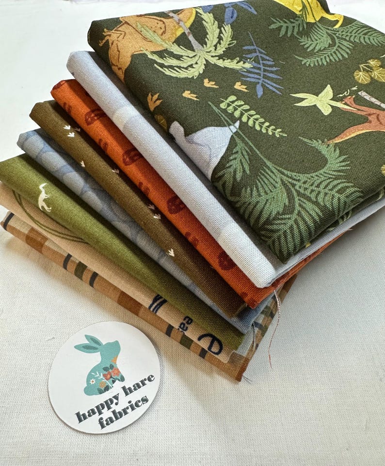 May include: A stack of six fabric squares with different patterns, including a green fabric with a jungle animal print, a blue fabric with a white animal print, and a brown fabric with a white footprint print. The fabric squares are stacked on top of each other and have a white background. The bottom fabric square has a brown and white striped pattern. A white circle with the text "happy hare fabrics" is in the lower left corner of the image.