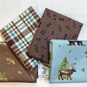 May include: A collection of folded fabric squares with various patterns. One features a blue and brown plaid, another has brown footprints, and a third has a forest scene with deer and bears. The fourth has a brown acorn and leaf pattern. A small sticker is visible.