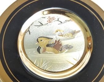 Könnte beinhalten: Ein dekorativer Teller mit einem schwarzen und goldenen Rand. Die Mitte des Tellers zeigt ein goldenes und silbernes Bild von zwei Enten in einem Teich mit einem Baumzweig darüber.