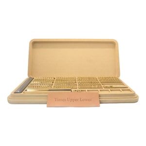 May include: Open case with a set of brass letter stamps for stamping leather. The case is made of a light brown material and has a label that reads "Times Upper Lower".