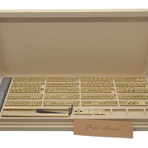 May include: A wooden box containing a set of brass letter stamps for stamping leather or metal. The box is open and the stamps are arranged in rows. The box also includes a metal tool and a small piece of leather with the text "Park Avenue" stamped on it.