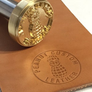 Puede incluir: Un sello de metal dorado con un diseño de cacahuete y el texto "Peanut Custom Leather" sobre una superficie de cuero marrón.