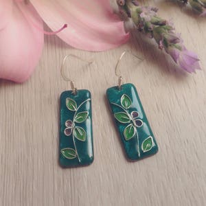 Puede incluir: Pendientes rectangulares de color azul turquesa con un alambre plateado y un diseño floral verde.