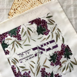 May include: White cloth with a hand-painted design of purple grapes and green leaves. The cloth has Hebrew text in purple. Two pieces of matzah bread are in the background. The cloth is likely used for a religious ceremony.