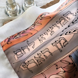 Può includere: Un tessuto di seta grigio e arancione con scrittura ebraica e spighe di grano. Il tessuto è piegato e ha uno sfondo bianco.