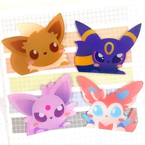 Puede incluir: Cuatro pegatinas de dibujos animados coloridos con Eevee, Umbreon, Espeon y Sylveon. La pegatina de Eevee es marrón, Umbreon es morado oscuro con detalles amarillos, Espeon es lavanda y Sylveon es rosa con detalles azules. Las pegatinas están sobre un fondo de cuadrícula.
