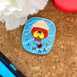 May include: A rectangular enamel pin with a cartoon beaver in a beige beanie and red shirt, floating in blue water. The pin has gold trim and the words "HEY, MAN" in white. The background is a light blue with white wave lines.