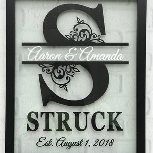 May include: Black framed shadow box with a black and white cutout of the letter "S" with a floral design. The text "Aaron & Amanda STRUCK Est. August 1, 2018" is below the letter. The bottom right corner of the frame has the text "Tam Tam's Cottage".