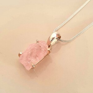 May include: A light pink, raw gemstone pendant on a silver chain. The pendant is a rectangular shape with a rough, natural texture. The silver chain is a delicate snake chain style.