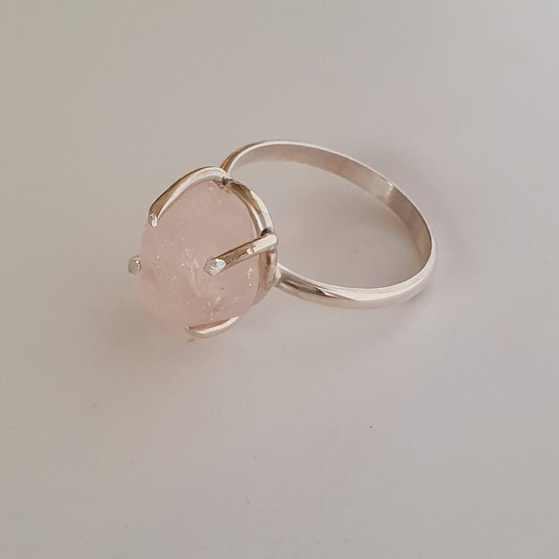 May include: A silver ring featuring a large, oval-shaped rose quartz gemstone. The pale pink stone is held in place by four prongs. The ring band is a simple, polished silver.