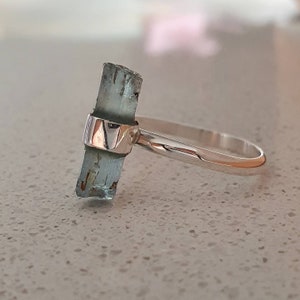 May include: A silver ring with a light blue aquamarine gemstone set in the center. The gemstone is a raw, unpolished crystal.