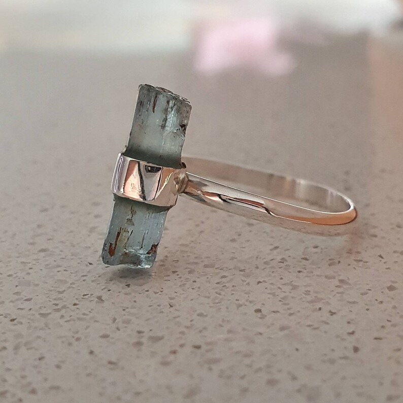 May include: A silver ring with a light blue aquamarine gemstone set in the center. The gemstone is a raw, unpolished crystal.