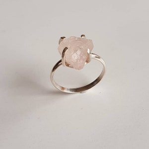 May include: A silver ring featuring a rough-cut, pale pink gemstone. The stone is held in place by four prongs, and the band is a simple, polished silver. The ring is set against a plain white background, highlighting the natural beauty of the stone.