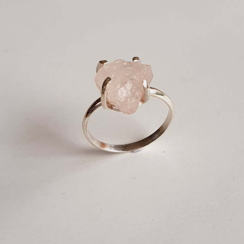 May include: A silver ring featuring a rough-cut, pale pink gemstone. The stone is held in place by four prongs, and the band is a simple, polished silver. The ring is set against a plain white background, highlighting the natural beauty of the stone.
