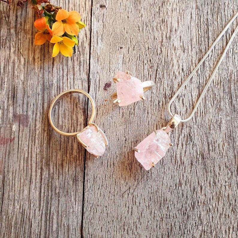 May include: A jewelry set featuring a gold ring, a pendant necklace, and a second ring, all with raw, light pink rose quartz stones. The jewelry is set against a rustic wooden background, with orange and yellow flowers in the upper left corner.