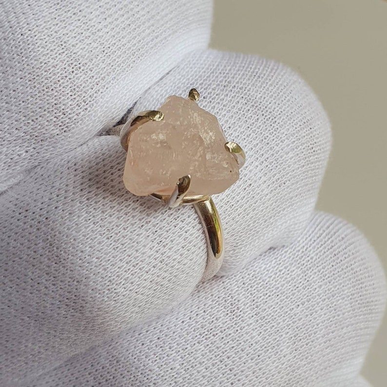 May include: A silver ring featuring a raw, light pink gemstone. The stone is irregularly shaped and held in place by four gold-colored prongs. The ring band is silver with a gold-colored accent near the stone.