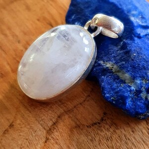 May include: A white oval moonstone pendant with a silver setting. The pendant is on a blue background.