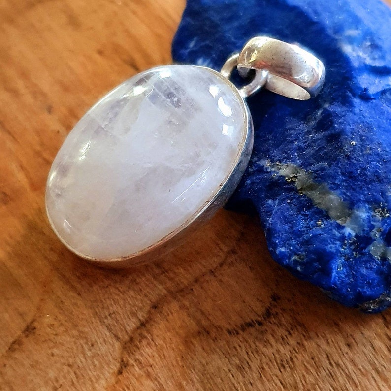 May include: A white oval moonstone pendant with a silver setting. The pendant is on a blue background.
