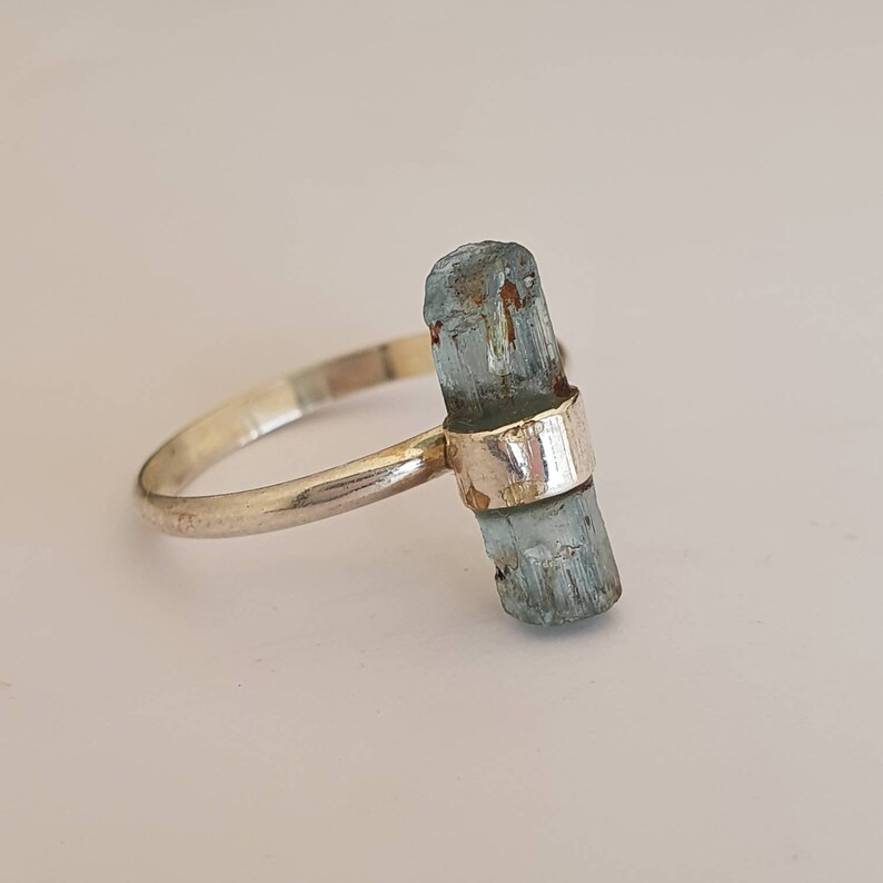 May include: A silver ring with a light blue aquamarine crystal set in the band. The crystal is a raw, natural piece with a rough texture.