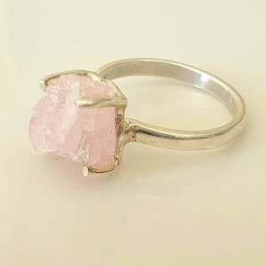 May include: A silver ring featuring a raw, light pink rose quartz gemstone. The stone is held in place by a four-prong setting. The ring band is a simple, polished silver.