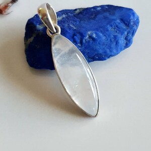 May include: A silver pendant with a clear, oval-shaped gemstone. The pendant is resting on a blue rock.