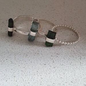 May include: Three silver rings with different colored gemstones. The rings are stacked on top of each other. The top ring has a dark green gemstone, the middle ring has a light blue gemstone, and the bottom ring has a black gemstone.