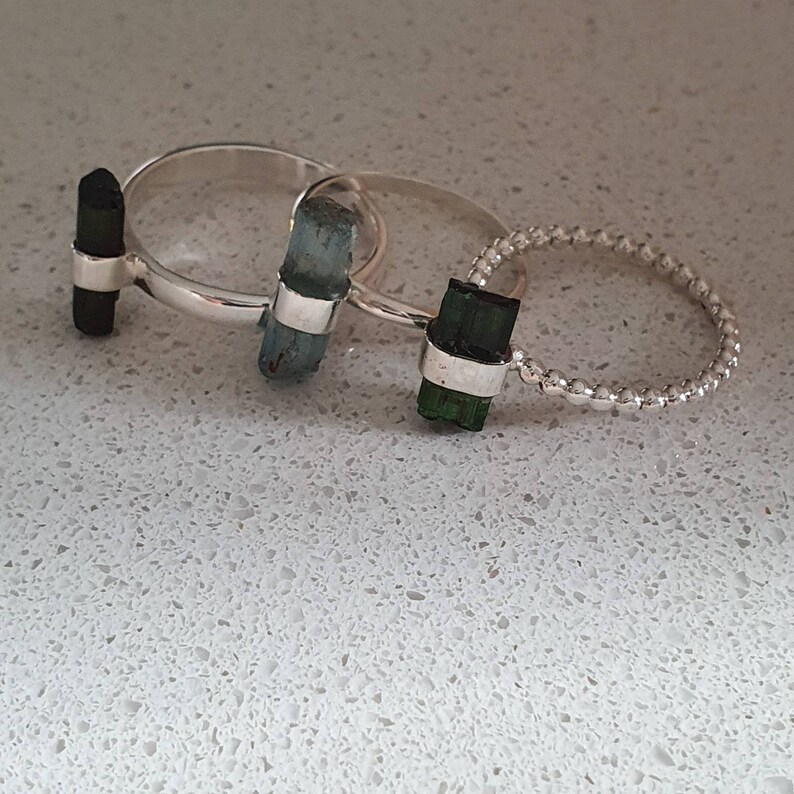 May include: Three silver rings with different colored gemstones. The rings are stacked on top of each other. The top ring has a dark green gemstone, the middle ring has a light blue gemstone, and the bottom ring has a black gemstone.