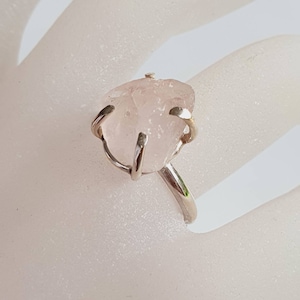 May include: A silver ring featuring a raw, light pink rose quartz gemstone. The stone is held in place by four silver prongs. The ring band is a simple, polished silver.