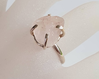 Puede incluir: Un anillo de plata con una gema de cuarzo rosa en bruto, de color rosa pálido. La piedra está sujeta por cuatro garras de plata. La banda del anillo es de plata pulida.