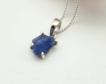May include: A silver necklace with a raw blue sapphire pendant. The sapphire is set in a silver bezel and has a rough, natural texture.