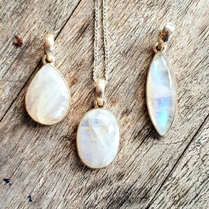 May include: Three silver pendants with moonstone stones. The stones are iridescent and have a white, blue, and gray color. The pendants are on a wooden surface.