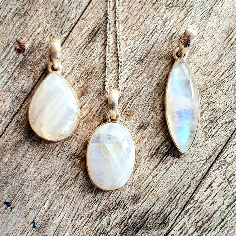 May include: Three silver pendants with moonstone stones. The stones are iridescent and have a white, blue, and gray color. The pendants are on a wooden surface.