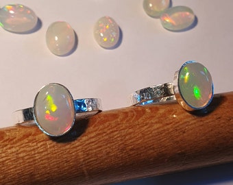 May include: Two silver rings with opal gemstones. The rings are on a wooden surface. The opals are oval shaped and have a white base with iridescent rainbow colors.