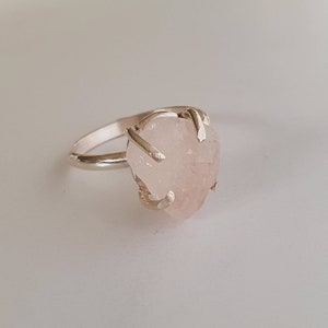 May include: A silver ring featuring a large, irregular-shaped rose quartz gemstone. The light pink stone is held in place by four silver prongs, creating a unique and natural look. The ring band is a simple, polished silver.