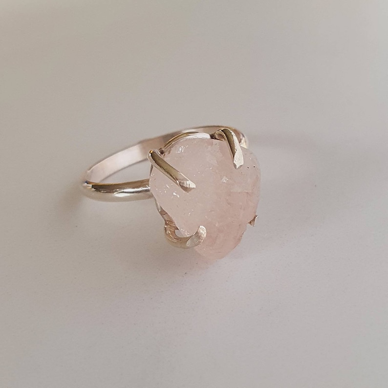 May include: A silver ring featuring a large, irregular-shaped rose quartz gemstone. The light pink stone is held in place by four silver prongs, creating a unique and natural look. The ring band is a simple, polished silver.