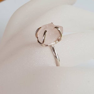 May include: A silver ring featuring a raw, light pink rose quartz gemstone. The stone is held in place by a silver wire setting, and the band is a simple, polished silver. The ring is a unique piece of jewelry.