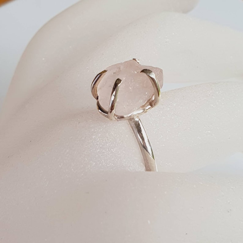 May include: A silver ring featuring a raw, light pink rose quartz gemstone. The stone is held in place by a silver wire setting, and the band is a simple, polished silver. The ring is a unique piece of jewelry.
